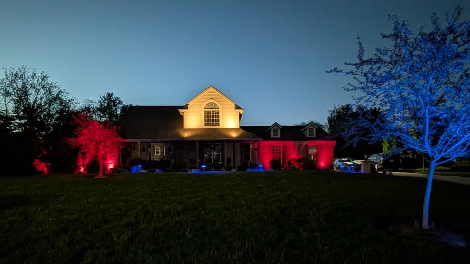 Home and trees lit with color-changing fixtures at dusk, the lawn dark in the foreground.