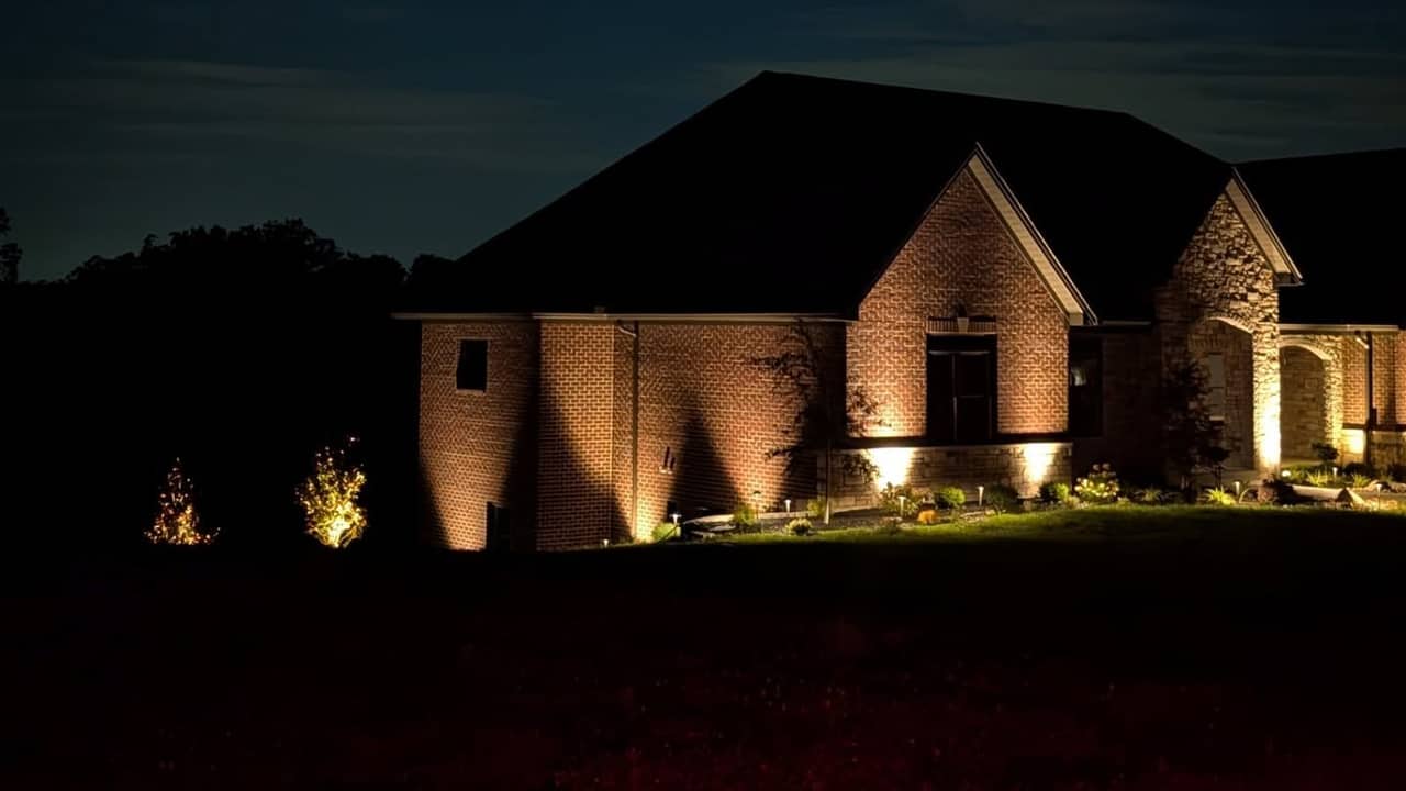 Two-story stone home with uplighting on the gables and warm light along the eaves after dark.