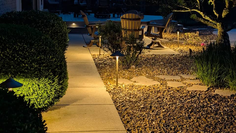Path lights leading along a walkway past a patio with chairs and a fire pit at dusk.