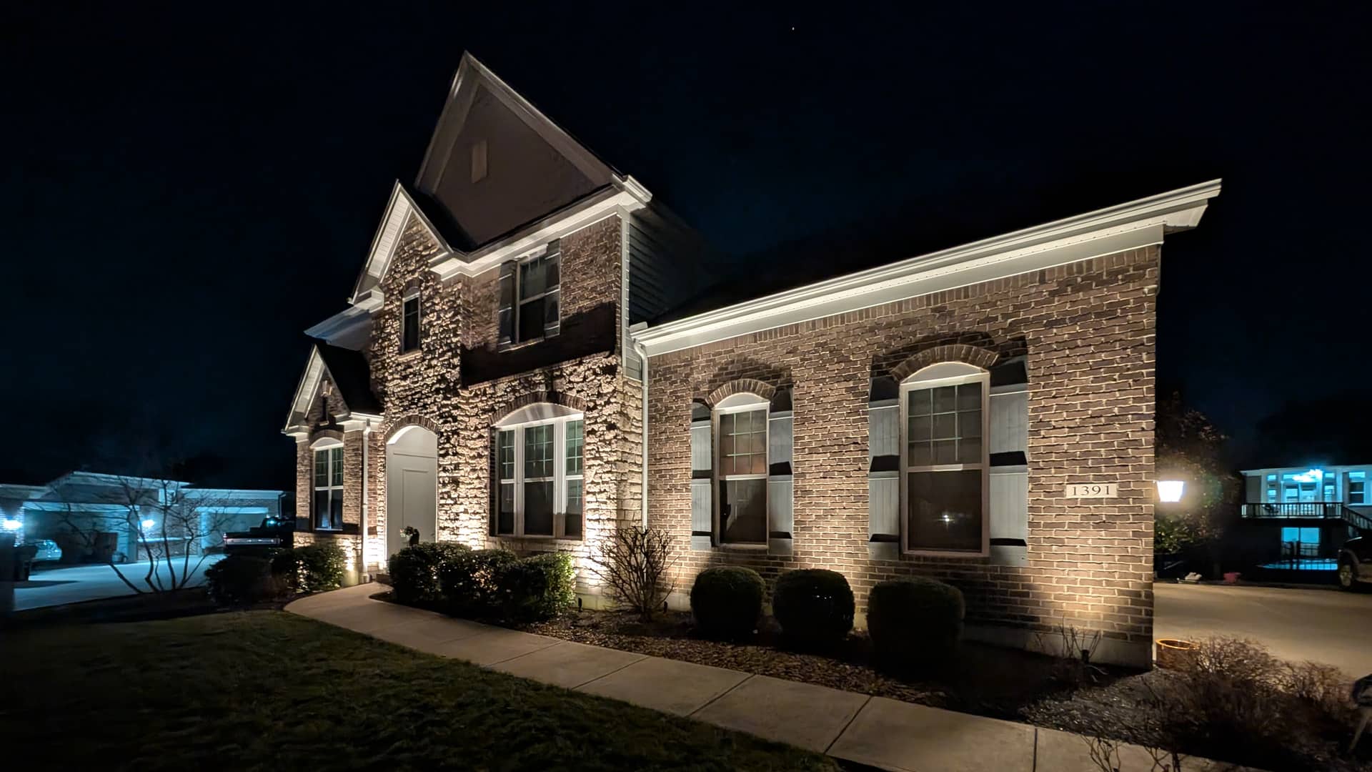 Brick and stone home lit by warm uplighting at night, with a lit walkway to the front door.